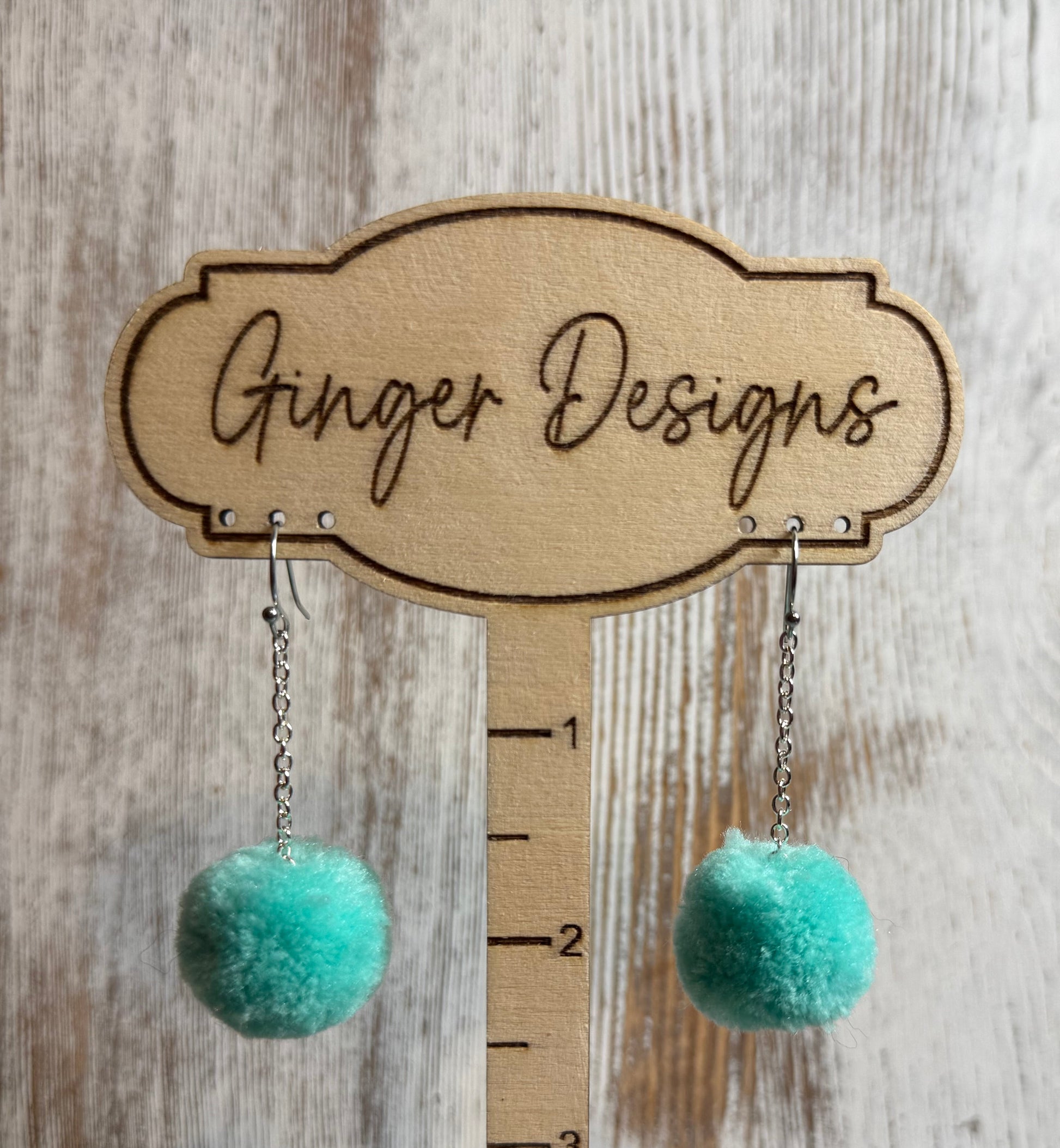 Turquoise pom-pom earrings on a wooden stand with 'Ginger Designs' branding.