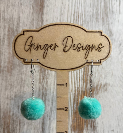 Turquoise pom-pom earrings on a wooden stand with 'Ginger Designs' branding.